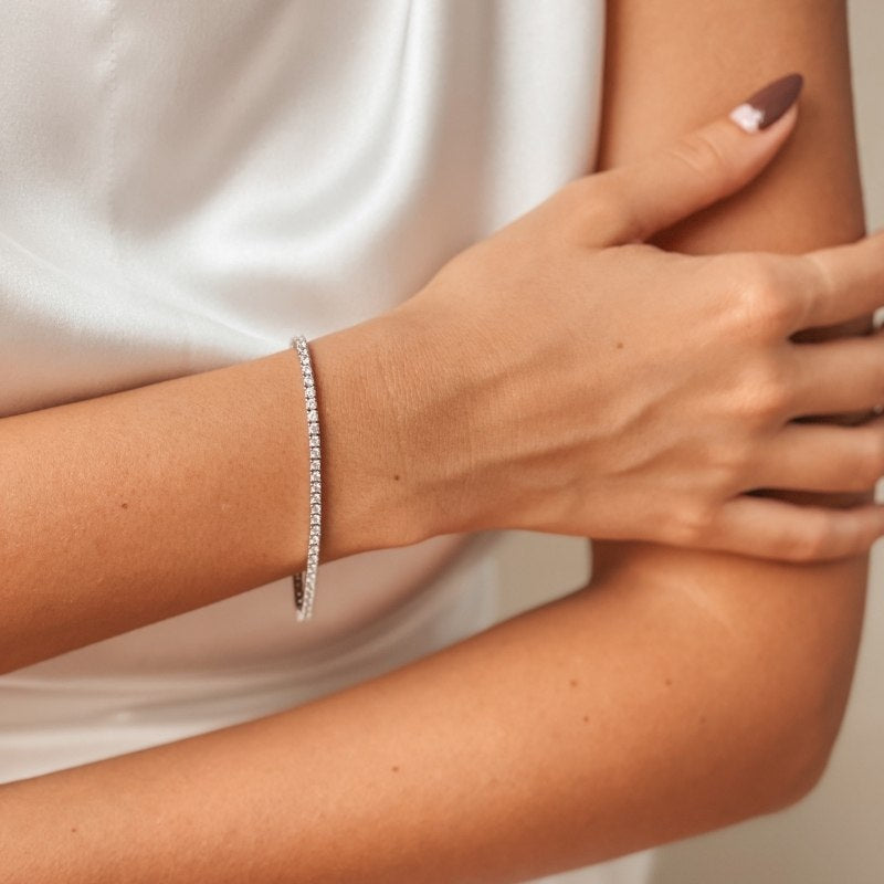 Close-up of a woman's arm adorned with a silver tennis bracelet featuring zirconia stones, set against a white satin background.
