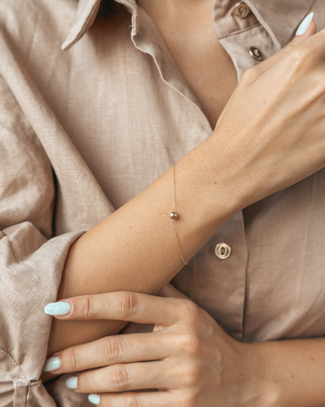 Close-up of a person wearing a delicate gold bracelet on their wrist, complemented by a beige shirt sleeve that enhances the elegance of the accessory.