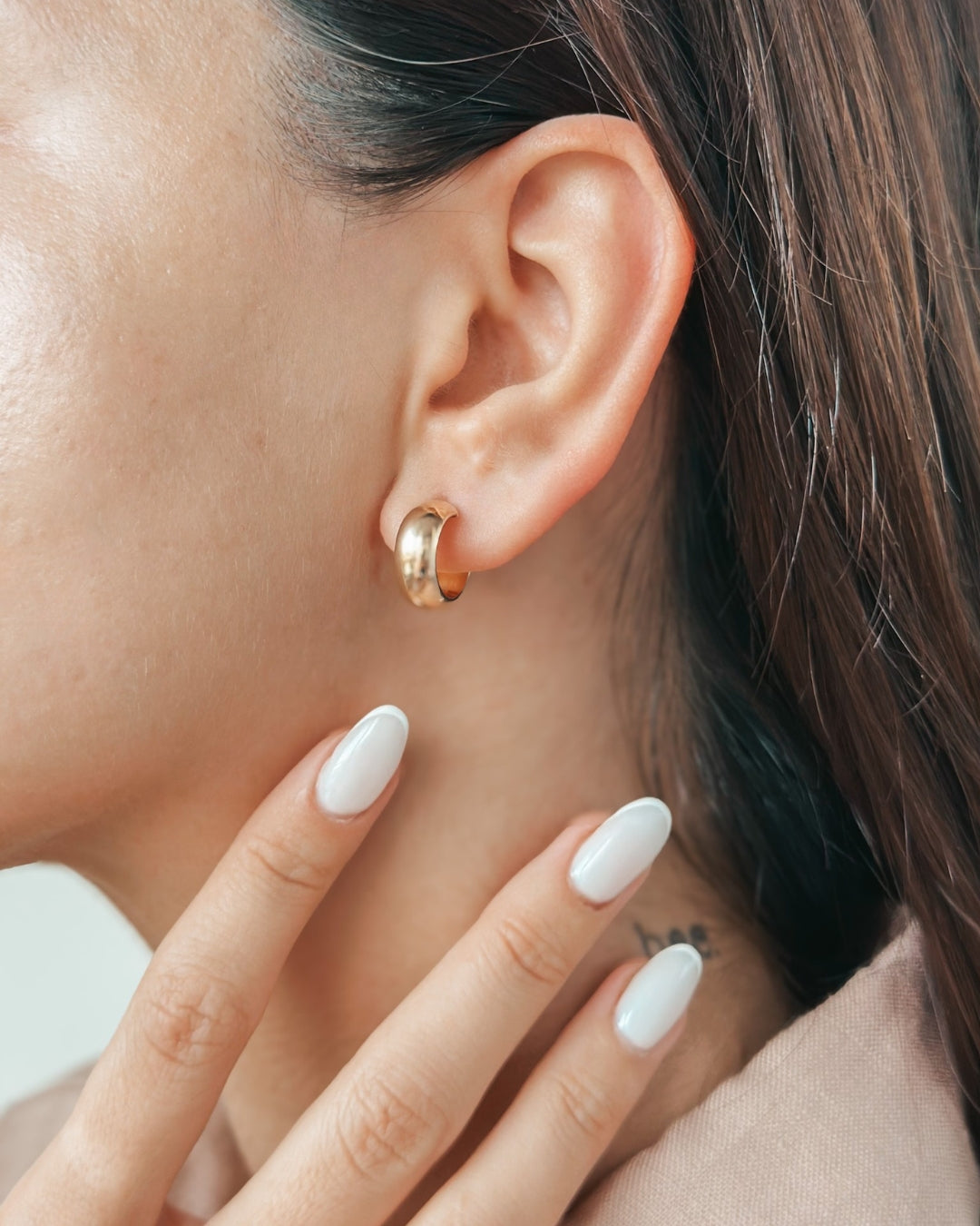 Close-up of a woman's ear adorned with a gold hoop earring, with manicured fingers gently resting on her neck.