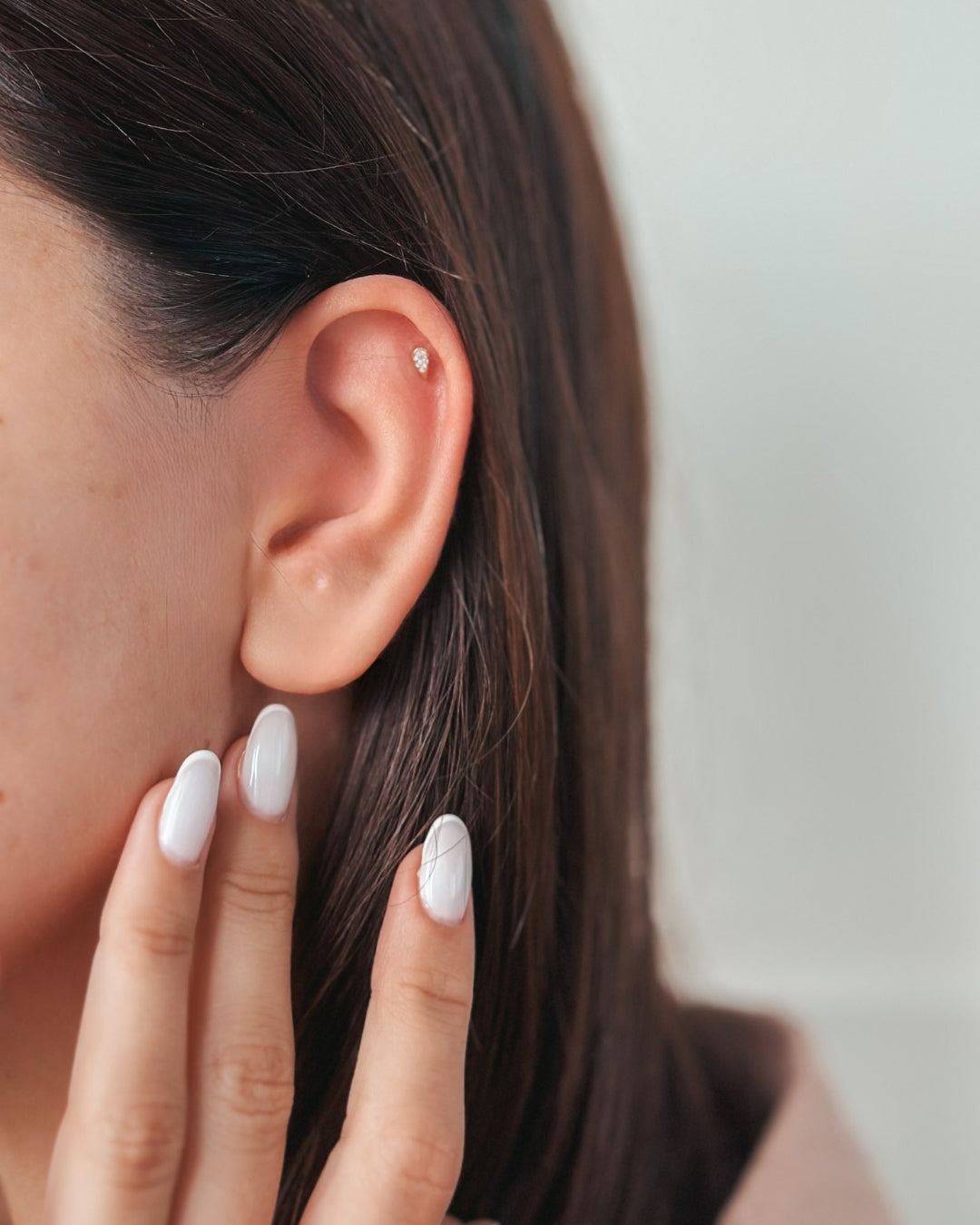 Close-up of an ear adorned with a small stud earring and fingers featuring white polished nails.