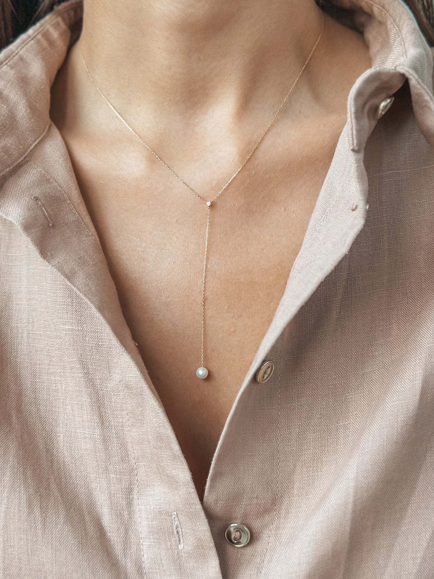 Close-up of a delicate gold necklace with a pearl pendant, worn over an open-collared shirt.