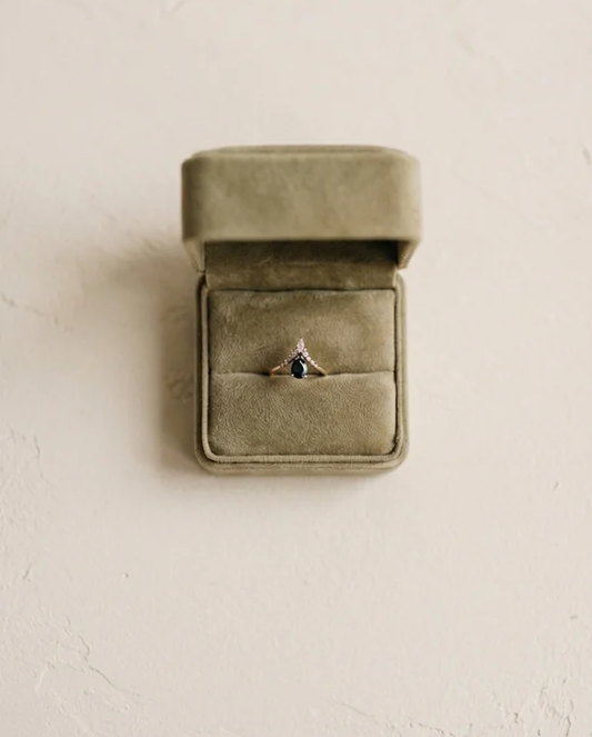 An open green velvet ring box containing a ring with a dark gemstone, placed on a light textured surface.