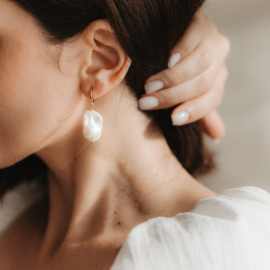 Close-up of an ear wearing a gold-plated 925 sterling silver hoop earring with a large white baroque pearl; a hand with a neutral manicure rests near the ear in soft natural light.