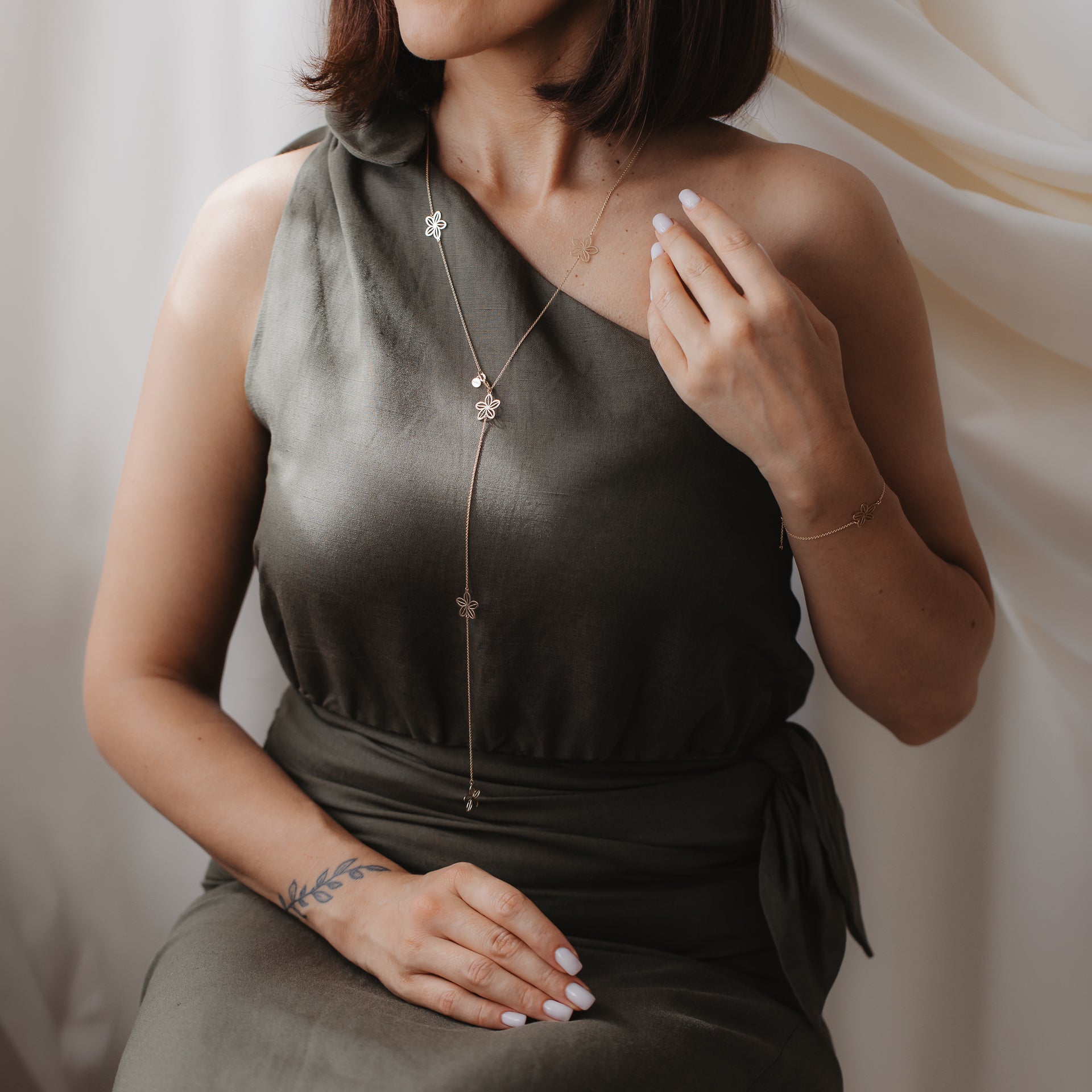 Close-up of a model wearing the Wildflower Maxi 14K yellow gold lariat necklace with floral charms over an olive one-shoulder dress.