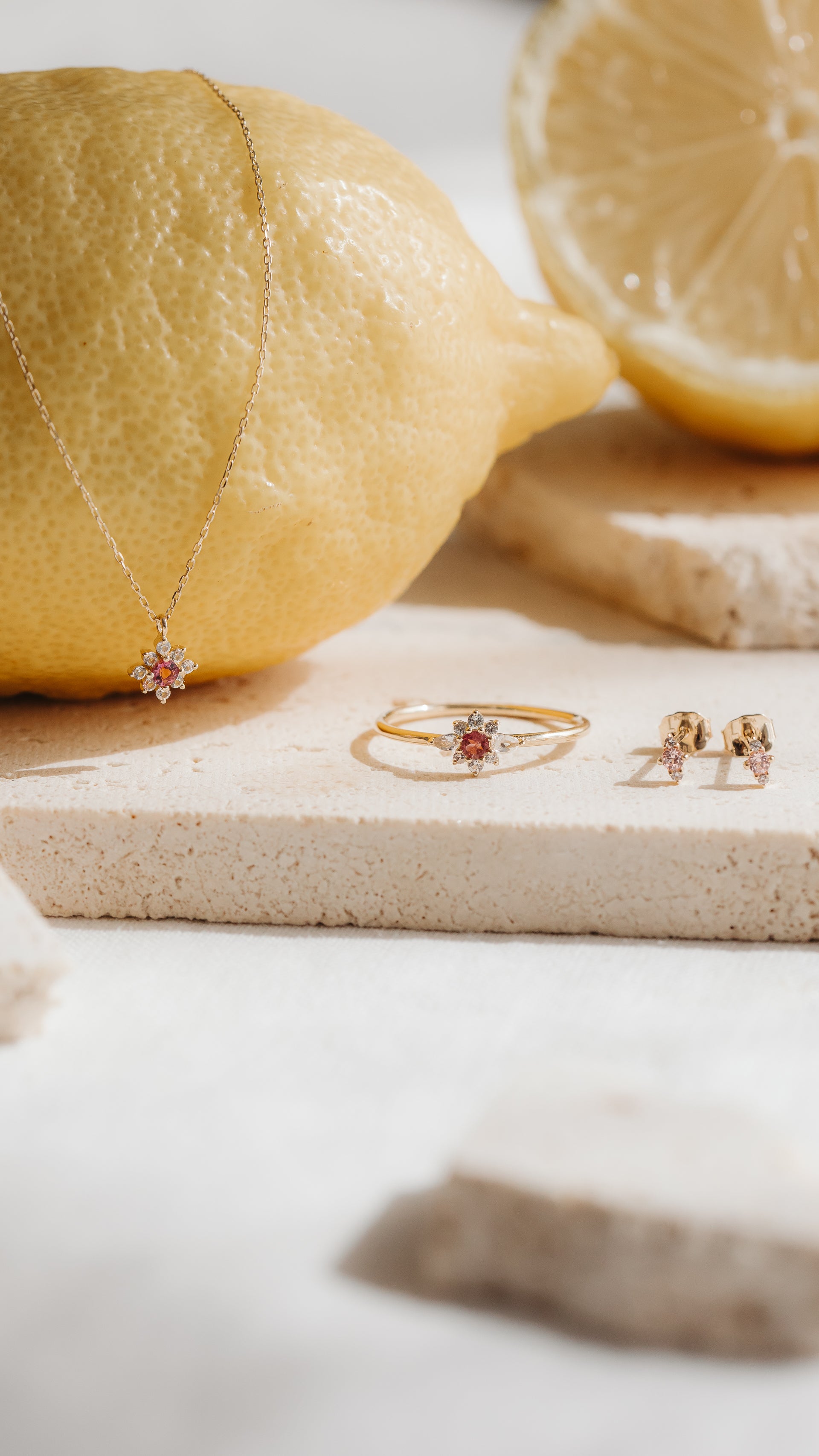 Flora 14K yellow gold stud earrings with pink morganite and white sapphires styled beside a matching ring and pendant on stone tiles with lemons.
