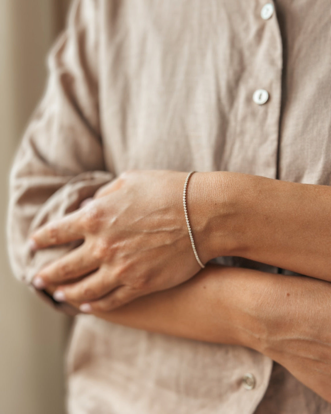 Close-up of an arm adorned with a delicate bracelet, crossed hands, wearing a linen shirt.