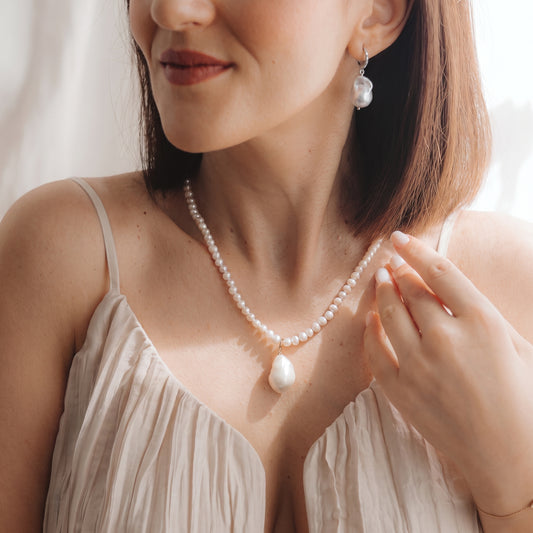 Close-up of a model wearing a rhodium-plated 925 silver baroque pearl drop earring with a small zirconia accent and a matching pearl necklace, hand near the collarbone in soft natural light.