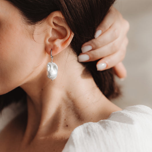 Close-up of an ear wearing a rhodium‑plated 925 silver hoop earring with a large, elegant baroque pearl drop, with a hand gently touching the hair.