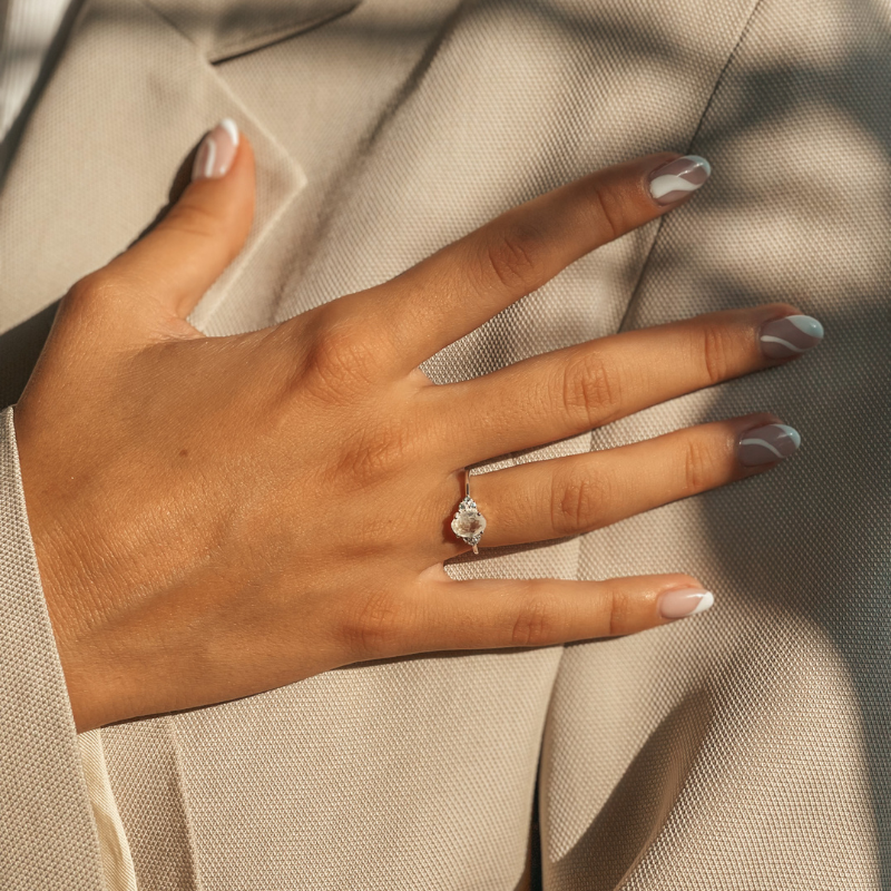 A hand with manicured nails adorned with a radiant silver ring featuring a moonstone, set against a textured beige fabric background.
