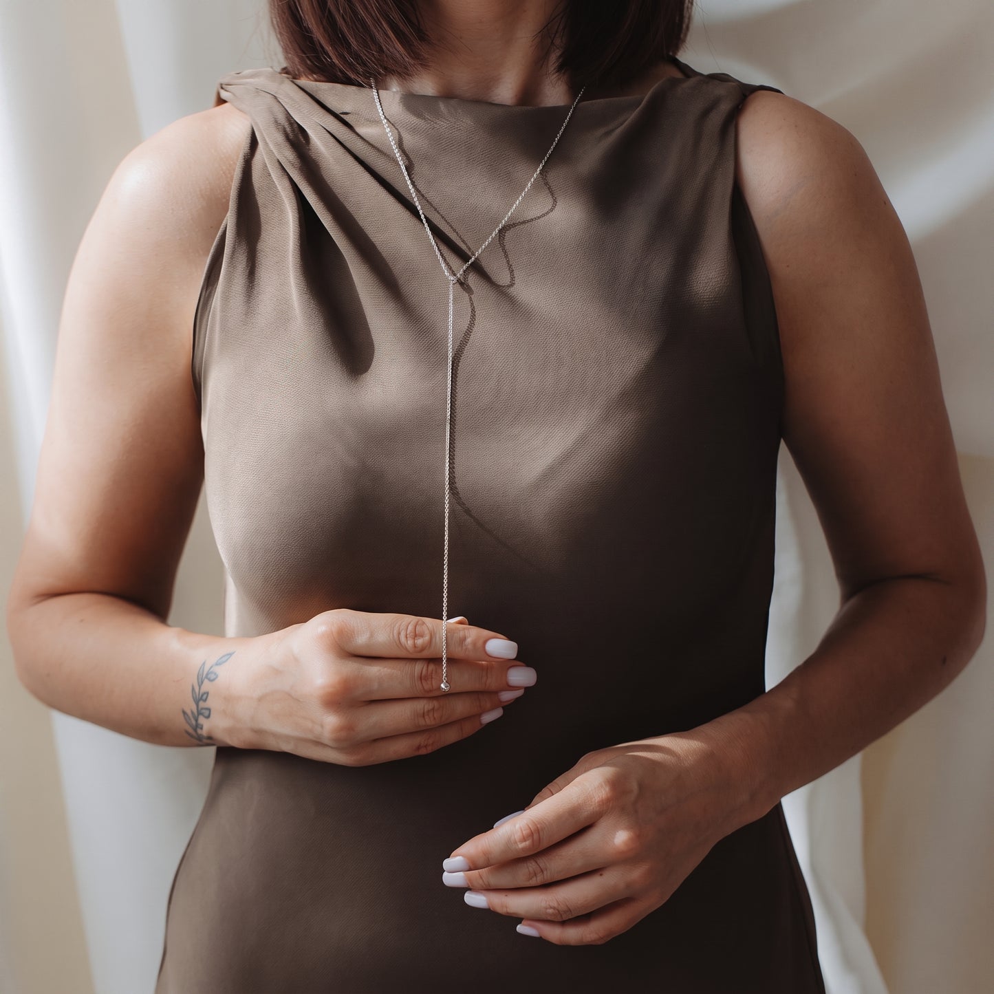 Close-up of a model wearing a 925 silver bola lariat necklace over a taupe sleeveless dress, lightly holding the drop chain.