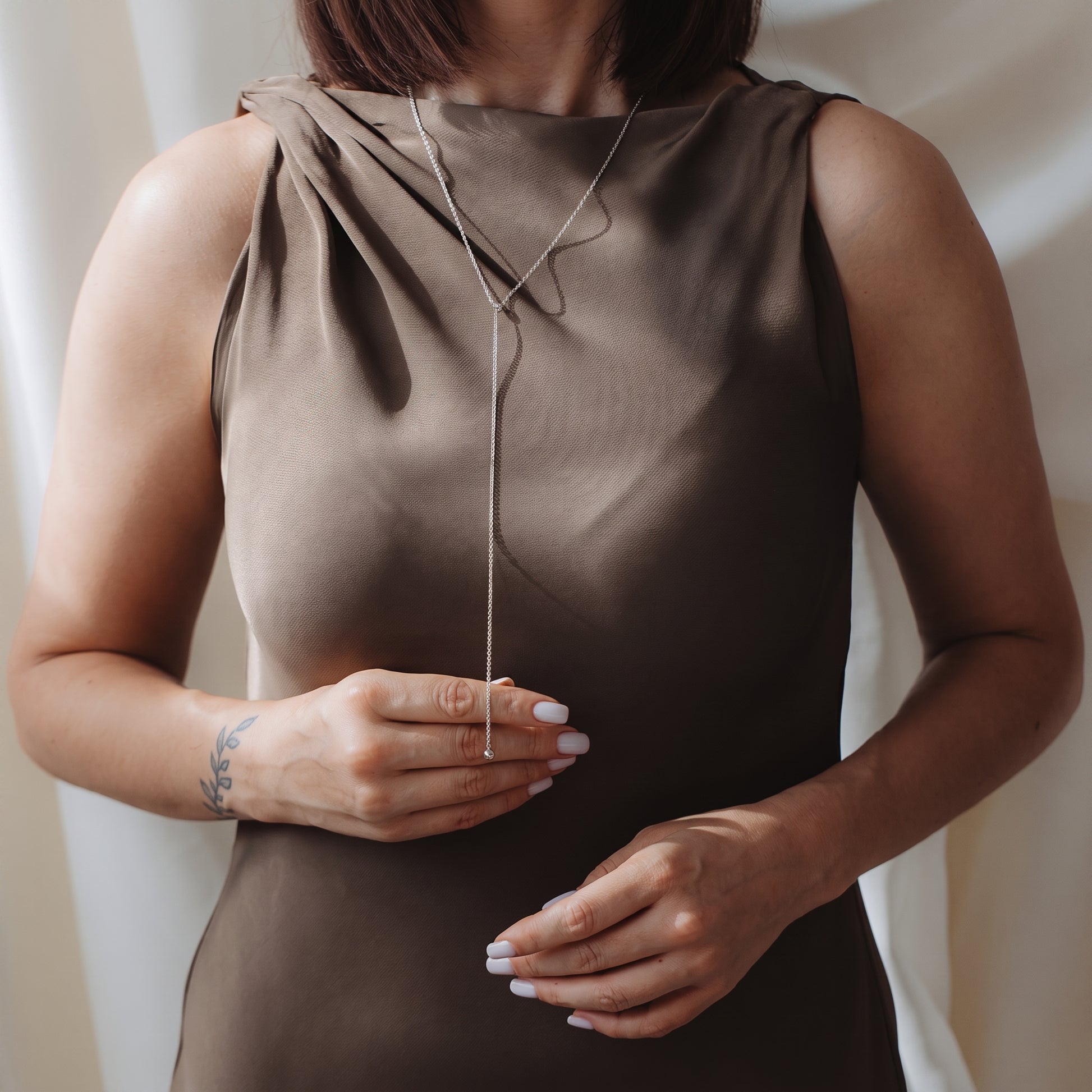 Close-up of a model wearing a 925 silver bola lariat necklace over a taupe sleeveless dress, lightly holding the drop chain.