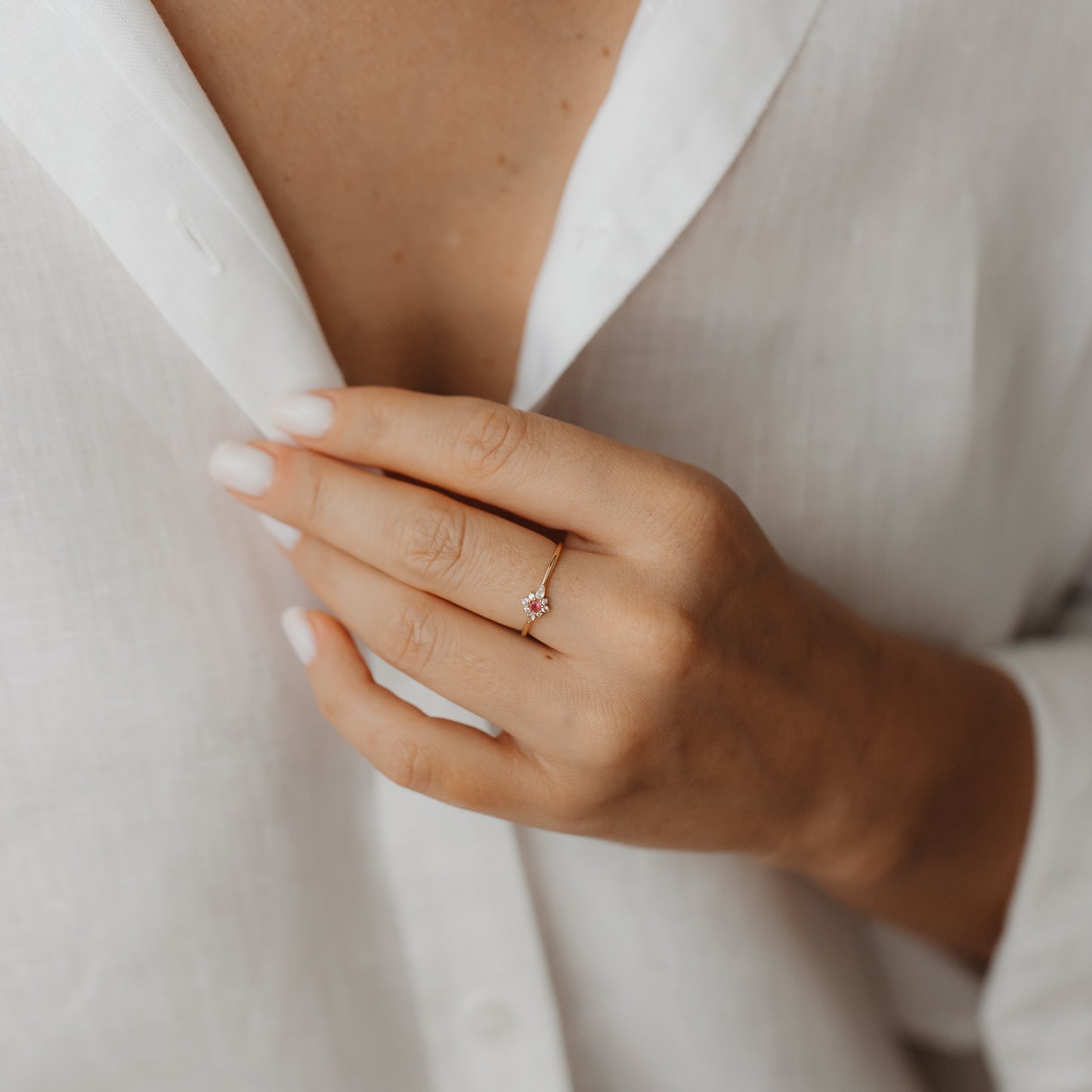 Close-up of a hand wearing the Flora 14K yellow gold ring with a white sapphire and pink tourmaline floral cluster, against a white linen shirt.