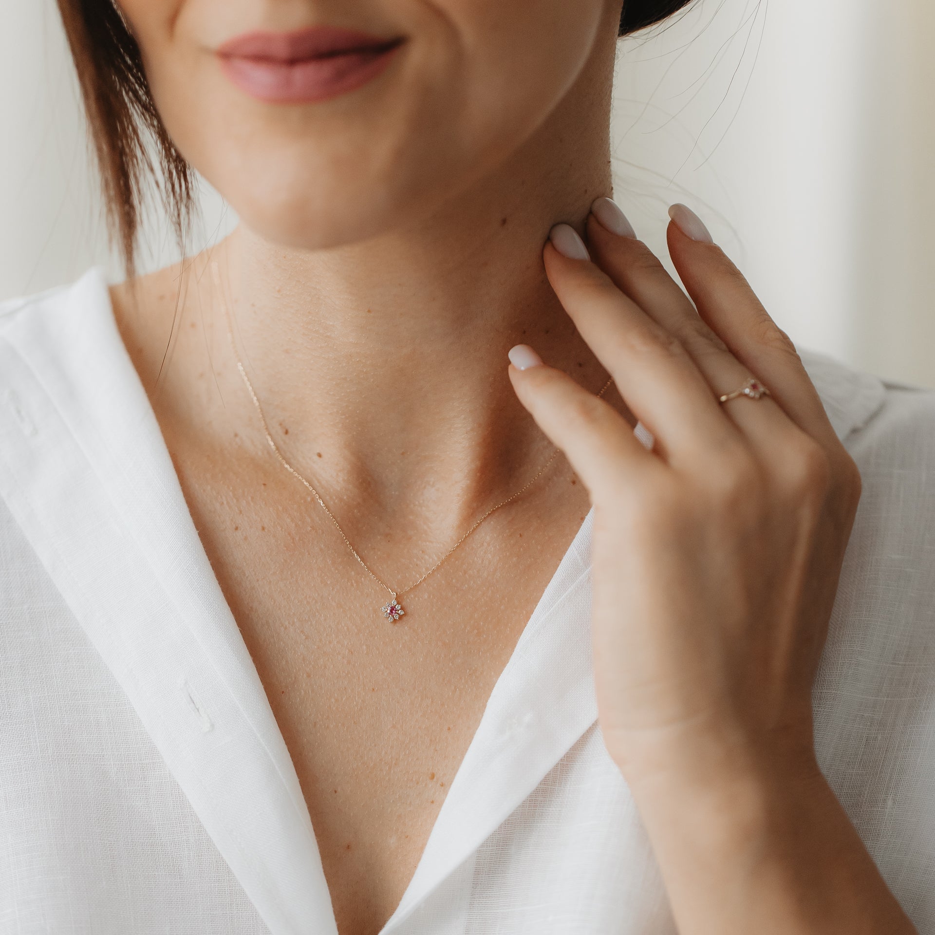 Close-up of the Flora 14k gold necklace featuring a tiny floral pendant set with pink tourmaline and white sapphires, with a hand touching the neck over a white blouse.