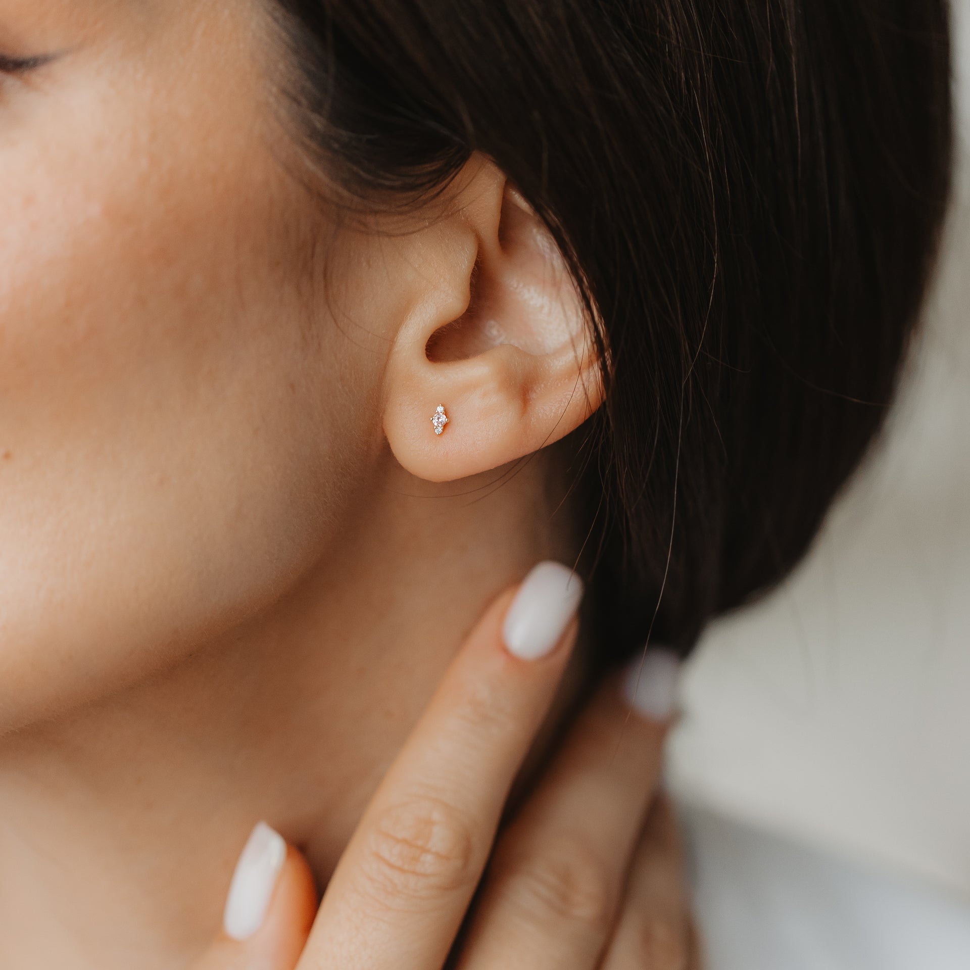Close-up of an ear wearing a tiny 14K yellow gold Flora stud earring with pink morganite and white sapphire, while a hand with a neutral manicure touches the neck.