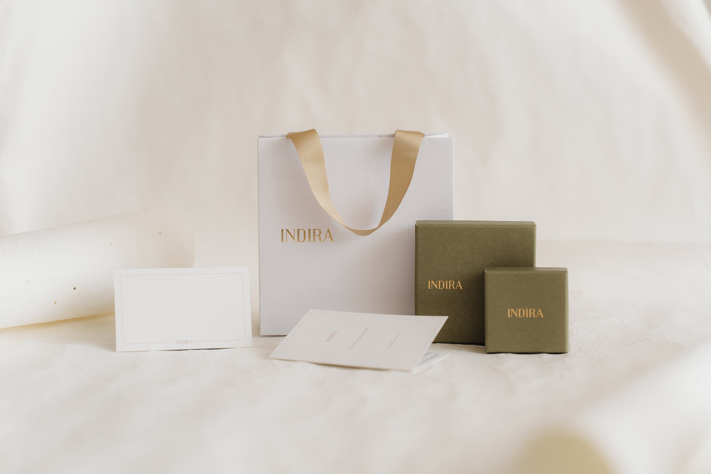 Minimal studio photo of INDIRA gift packaging set: a white bag with gold ribbon handles, two olive-green jewelry boxes, and blank cards on a soft ivory background.