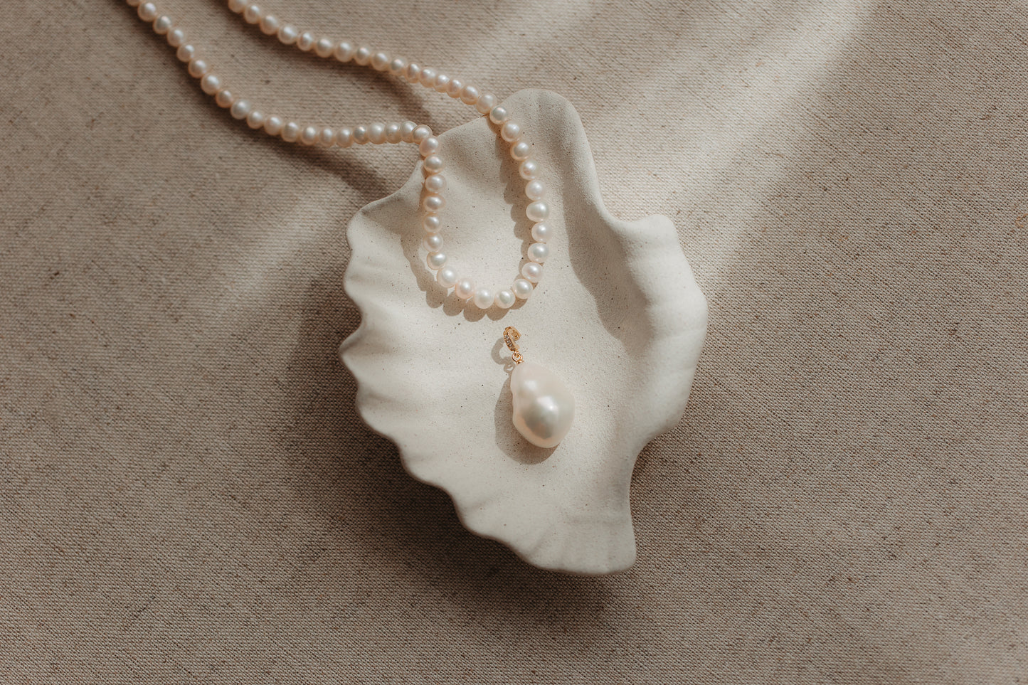 Baroque pearl pendant with a gold‑plated silver bail resting in a scalloped ceramic shell dish, with a pearl strand on a beige linen background.