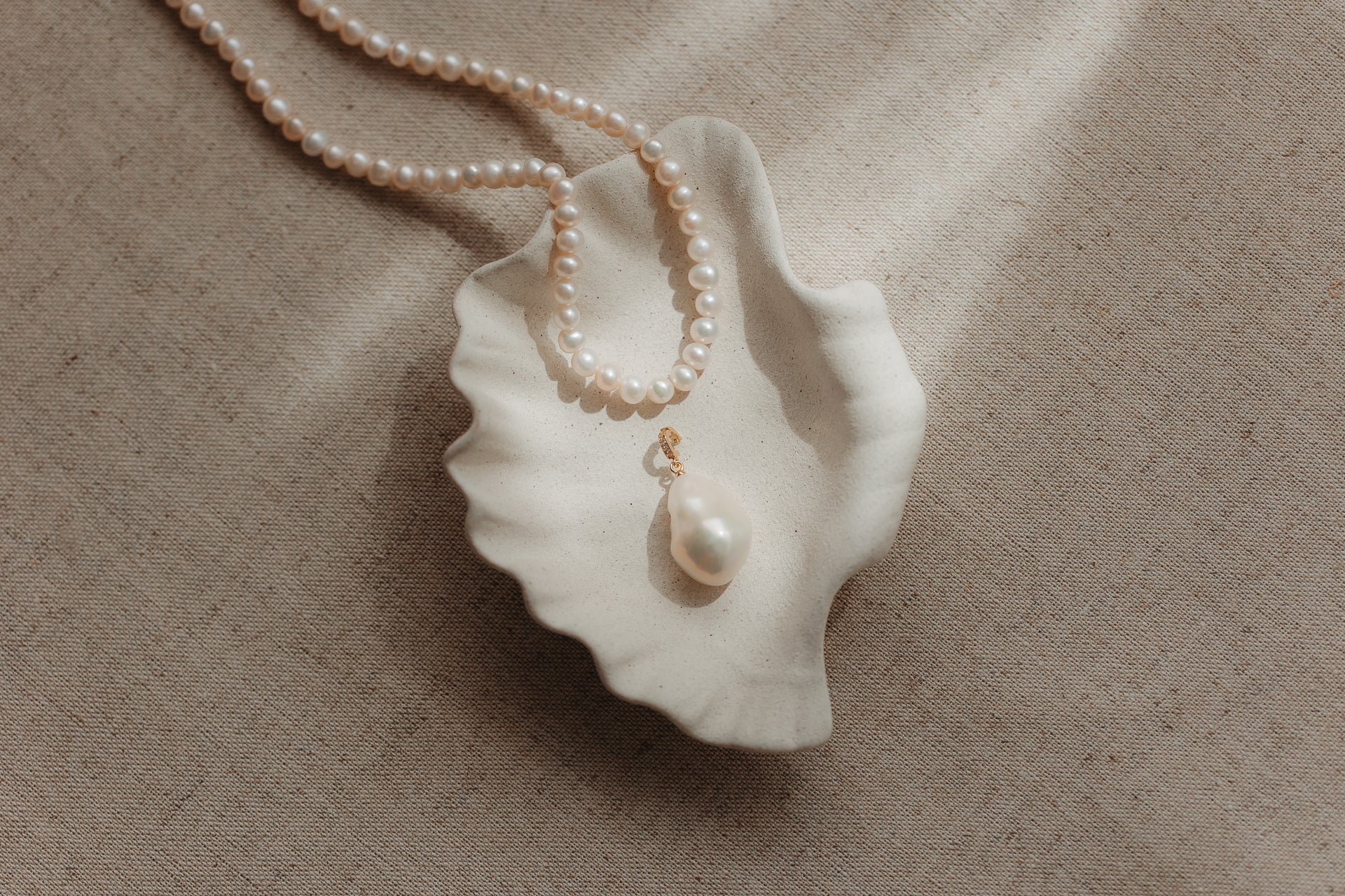 Baroque pearl pendant with a gold‑plated silver bail resting in a scalloped ceramic shell dish, with a pearl strand on a beige linen background.