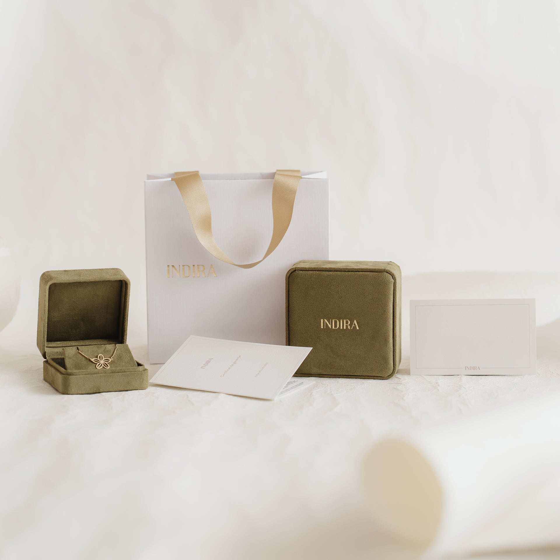 Indira-branded gift packaging set featuring a white bag with gold ribbon handles, olive velvet jewelry boxes (one open), and cards arranged on a soft neutral background.