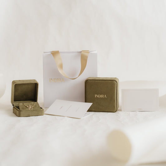 Indira-branded gift packaging set featuring a white bag with gold ribbon handles, olive velvet jewelry boxes (one open), and cards arranged on a soft neutral background.