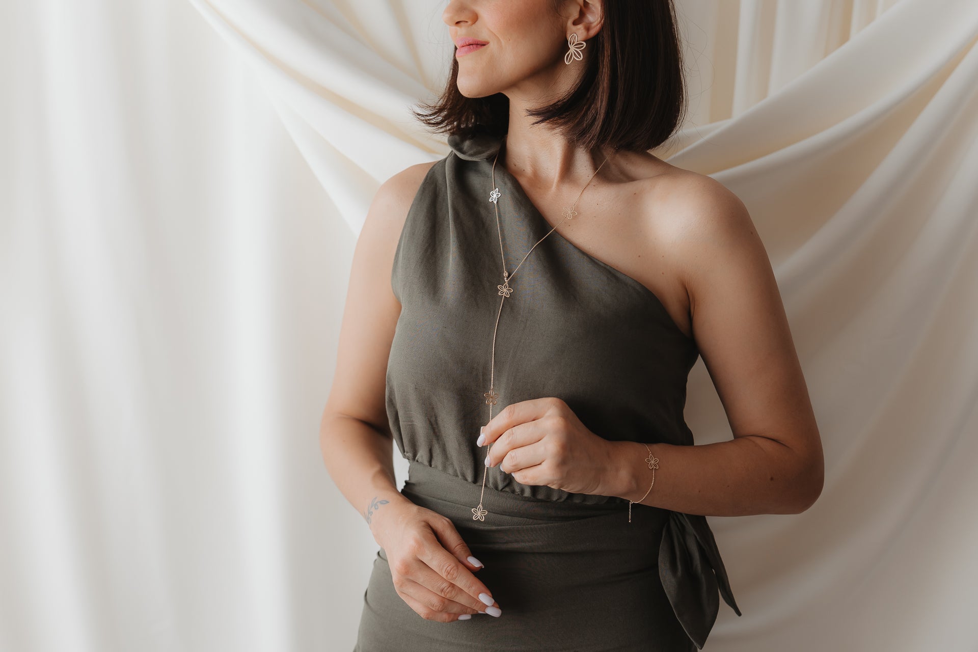 Close-up of a model wearing the Wildflower Maxi 14K yellow gold lariat necklace, featuring delicate flower charms, styled over an olive one-shoulder dress against a soft, draped cream backdrop.