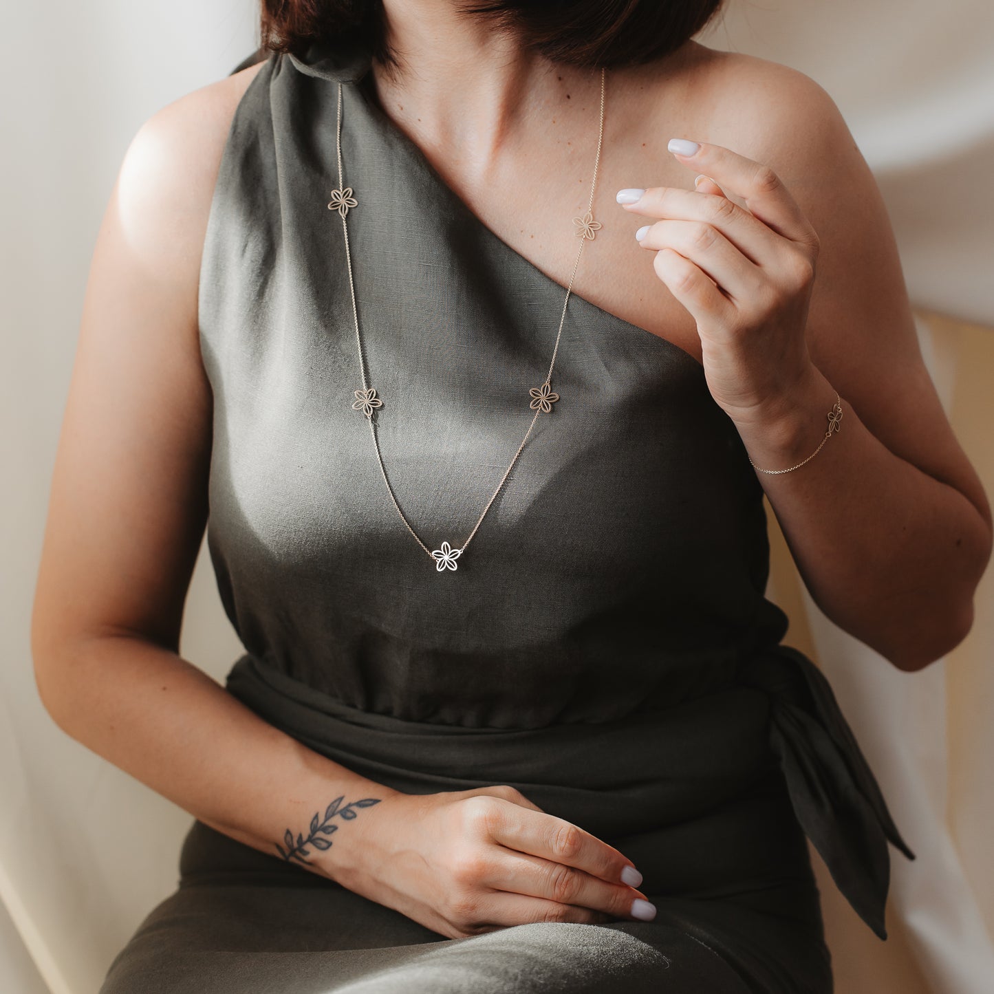 Close-up of a model in an olive one-shoulder dress wearing the Wildflower Maxi 14K yellow gold station necklace with small flower charms, shown in soft indoor light with a matching bracelet.