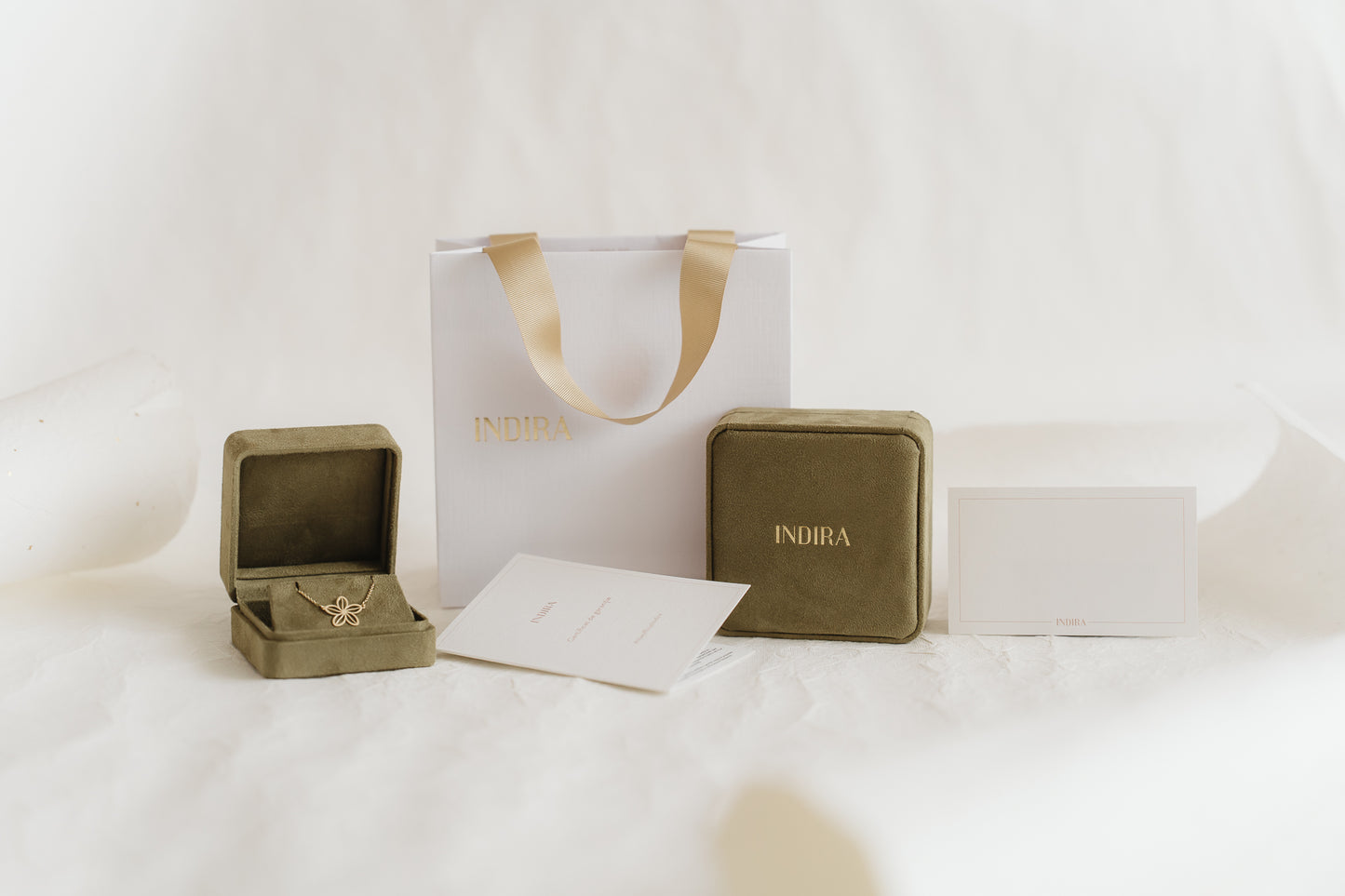 Indira gift packaging set featuring an elegant white shopping bag with gold ribbon handles, two olive velvet jewelry boxes (one open revealing a gold flower pendant), and branded cards on a soft white studio background.