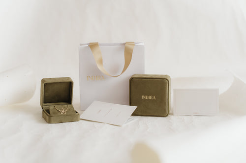 Indira gift packaging set featuring an elegant white shopping bag with gold ribbon handles, two olive velvet jewelry boxes (one open revealing a gold flower pendant), and branded cards on a soft white studio background.