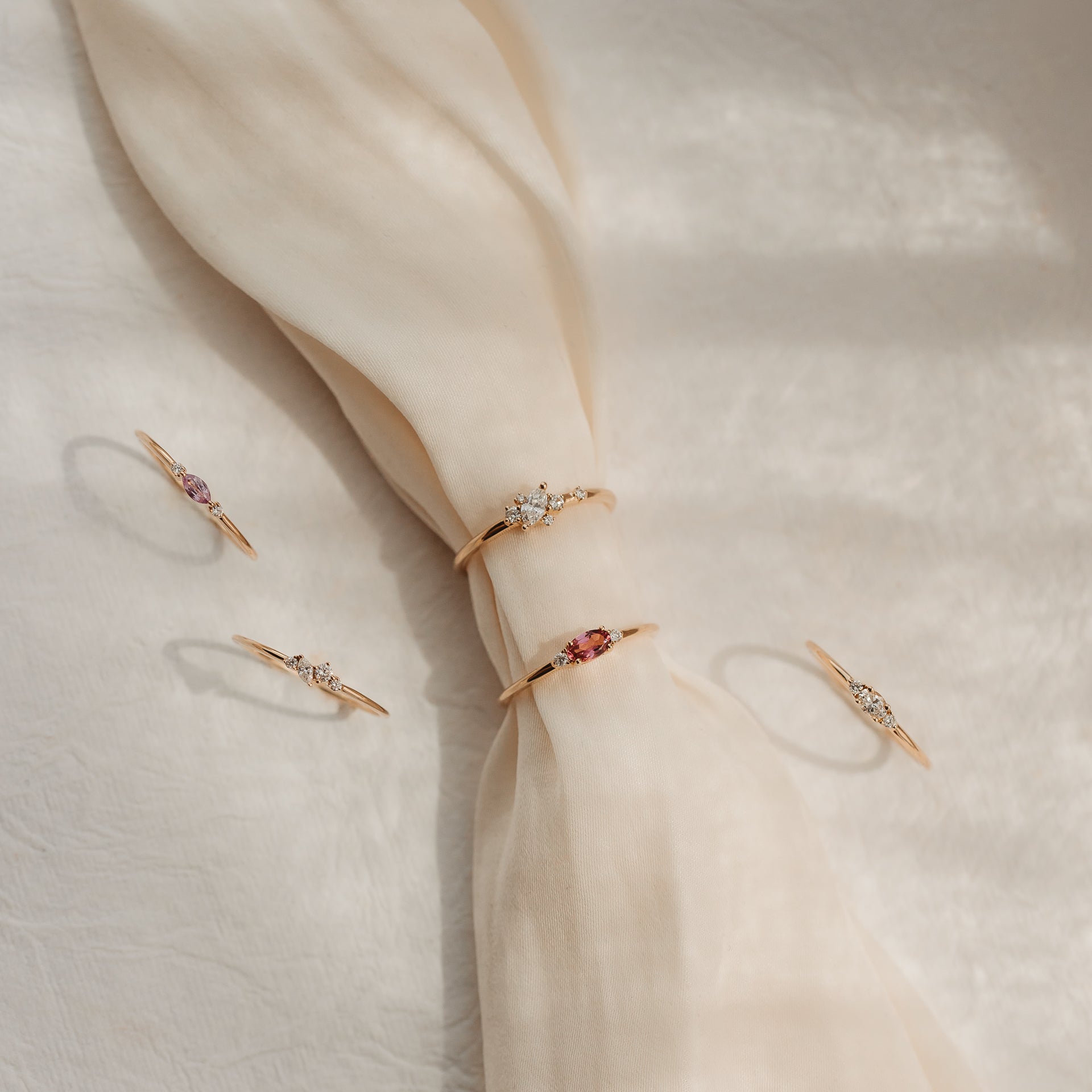 Five delicate 14k yellow-gold rings with diamonds and pink gemstones styled on a knotted cream silk fabric in soft sunlight.