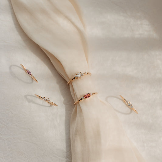 Five delicate 14k yellow-gold rings with diamonds and pink gemstones styled on a knotted cream silk fabric in soft sunlight.