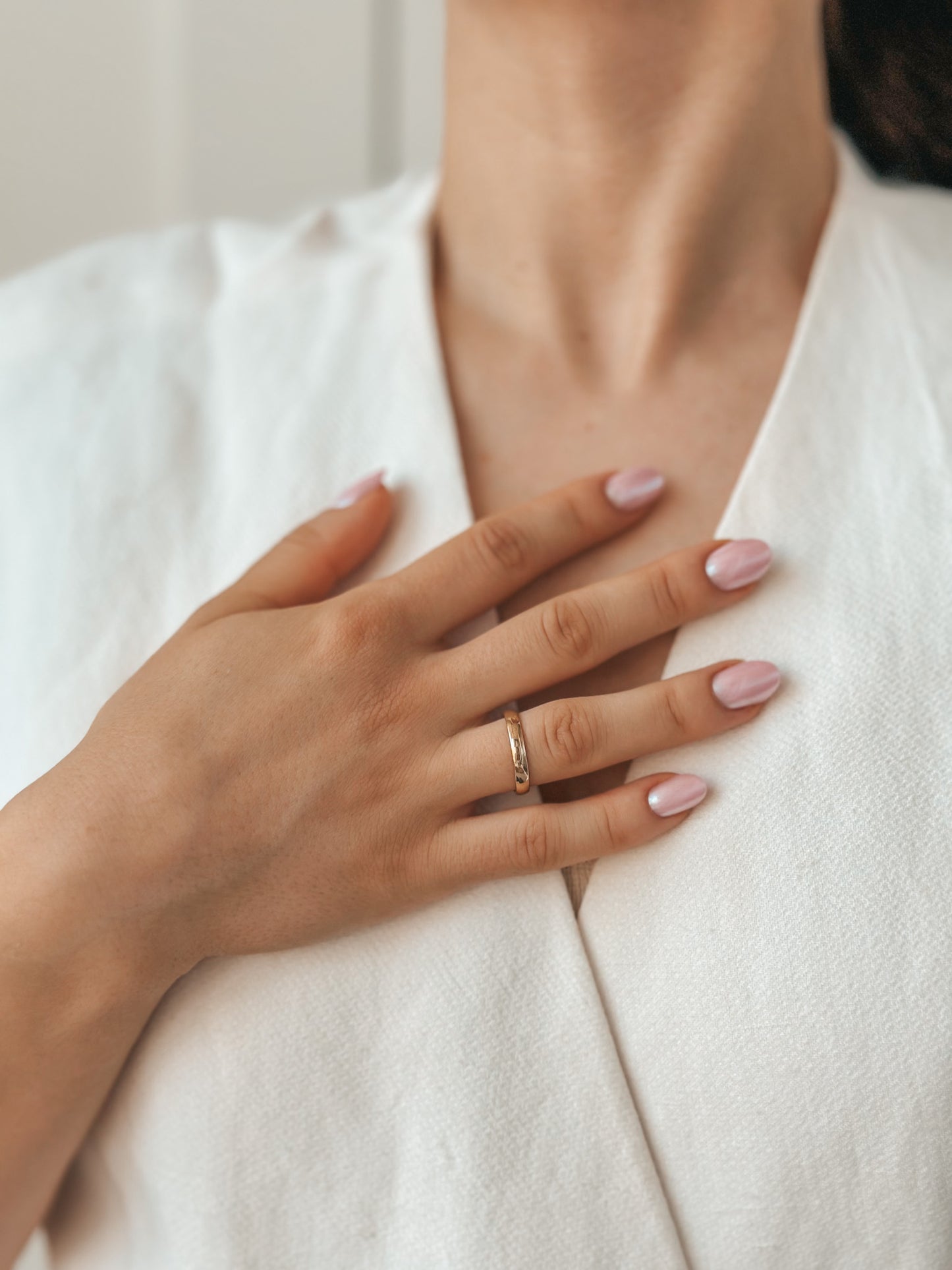 Close-up of a hand with a pink-polished manicure resting on the chest, adorned with a gold wedding band and wearing a white textured garment.