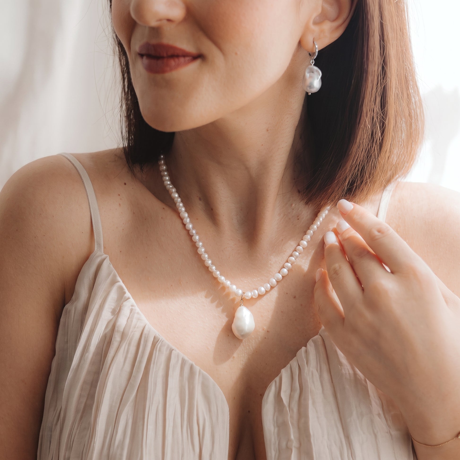 Close-up of a model wearing a rhodium-plated 925 silver baroque pearl drop earring with a small zirconia accent and a matching pearl necklace, hand near the collarbone in soft natural light.
