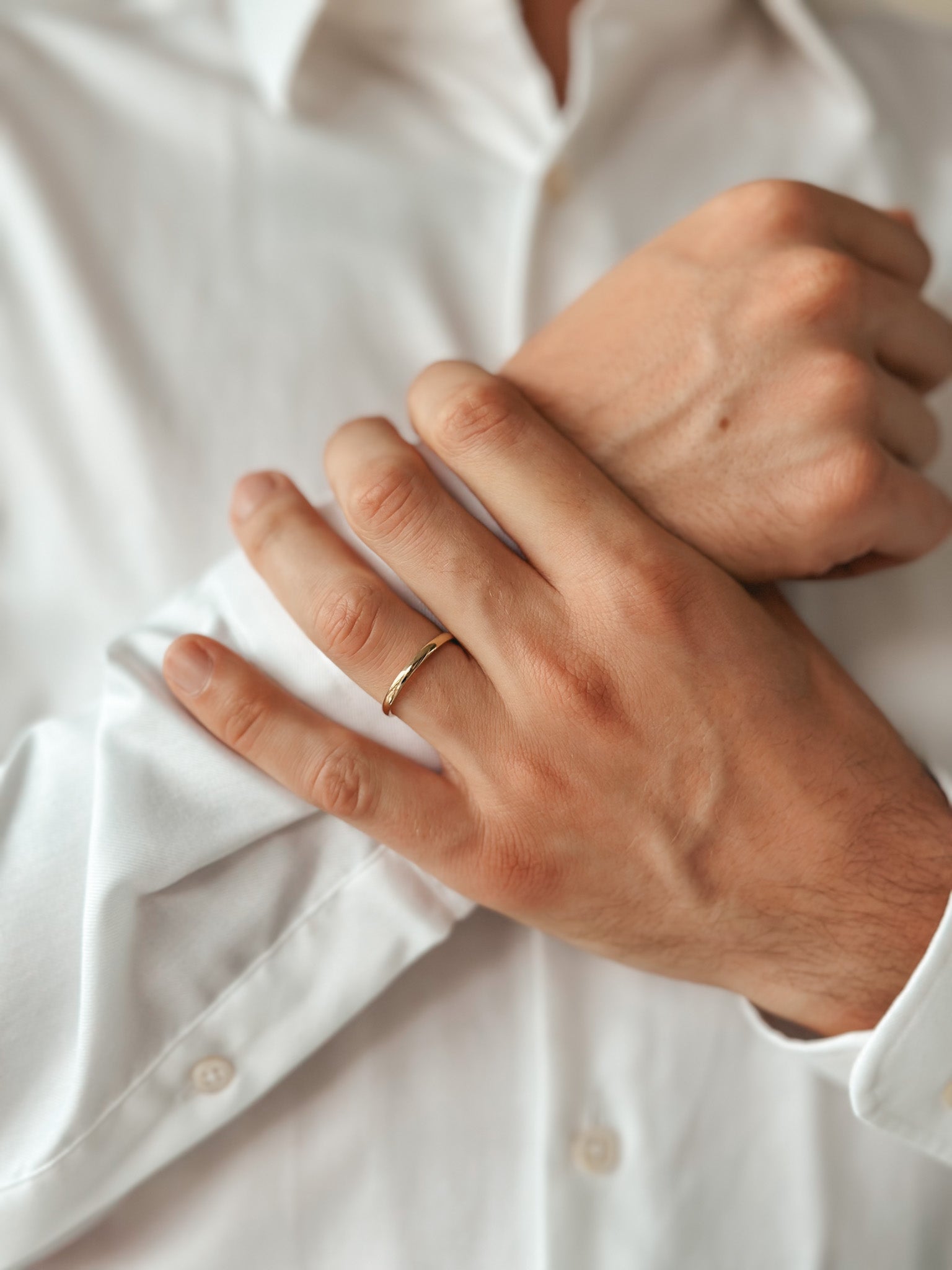 Close-up of a person wearing a thin, rounded gold wedding band on their hand over a white shirt.