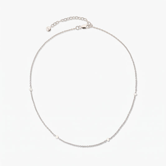 Delicate silver chain necklace with tiny white pearl stations, arranged in a circle on a white background, featuring an adjustable extender and lobster clasp.